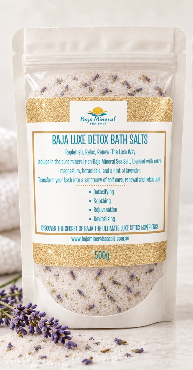 A stand-up pouch of Baja Luxe Detox Bath Salts by Baja Mineral Spa, filled with pale lavender bath salts, shown on a light surface with lavender sprigs at the base.