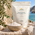 A white stand-up pouch of Baja Mineral Sea Salt stands on a stone surface outdoors, with a bowl of coarse white salt in front and a beach scene with blue water and sky in the background. The pouch label reads Baja Mineral Sea Salt and Baja X Coarse.