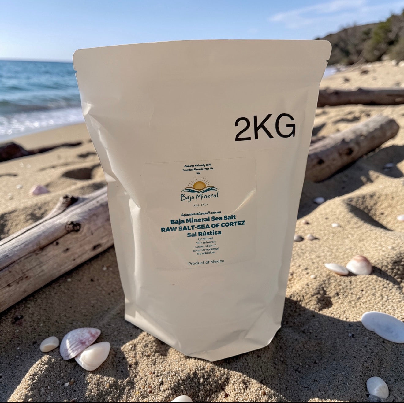 A 2KG stand-up pouch of Baja Mineral Sea Salt on a sandy beach with shells and driftwood, with the ocean and sky in the background.