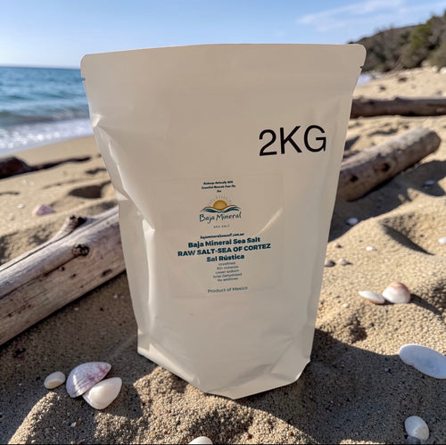 A 2KG stand-up pouch of Baja Mineral Sea Salt on a sandy beach with shells and driftwood, with the ocean and sky in the background.