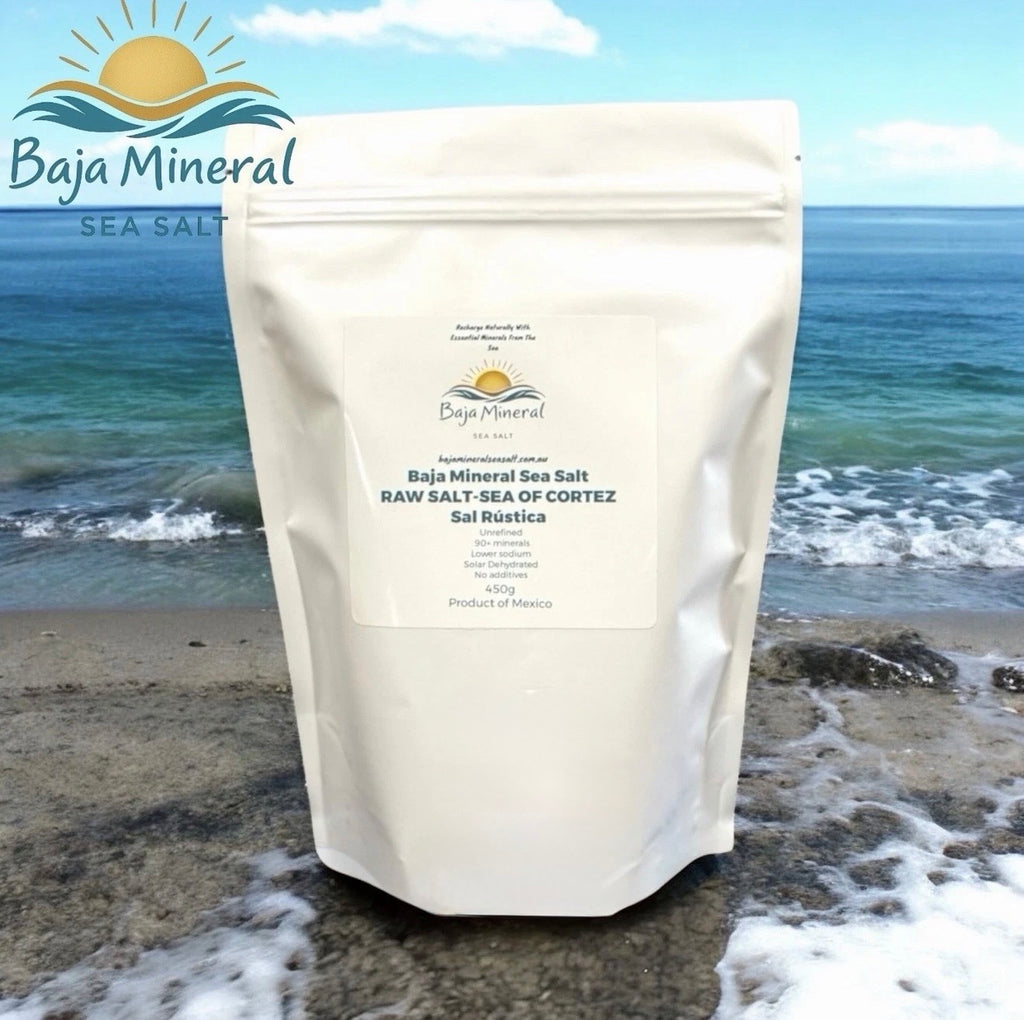A stand-up pouch of Baja Mineral Sea Salt standing on a beach with ocean waves and shoreline in the background. The pouch is off-white with a large front label reading Baja Mineral Sea Salt and product details.