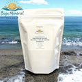 A stand-up pouch of Baja Mineral Sea Salt standing on a beach with ocean waves and shoreline in the background. The pouch is off-white with a large front label reading Baja Mineral Sea Salt and product details.