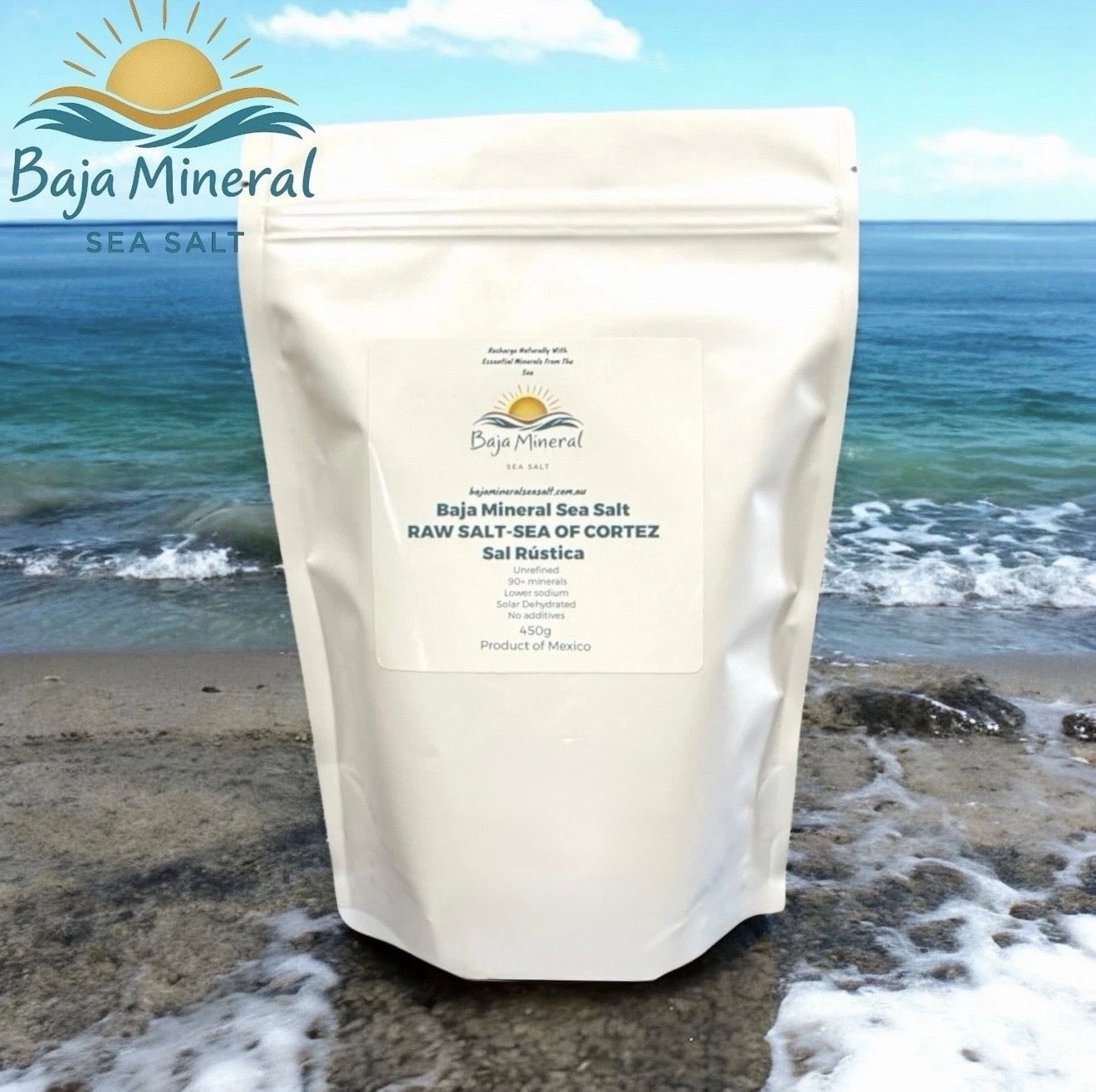 A stand-up pouch of Baja Mineral Sea Salt standing on a beach with ocean waves and shoreline in the background. The pouch is off-white with a large front label reading Baja Mineral Sea Salt and product details.