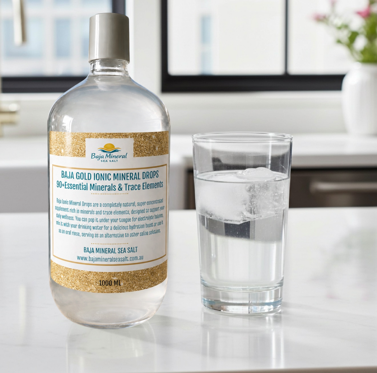 A clear bottle of Baja Gold Ionic Mineral Drops with a white cap and a cream label, standing on a white countertop next to a clear glass of water in a bright kitchen setting.